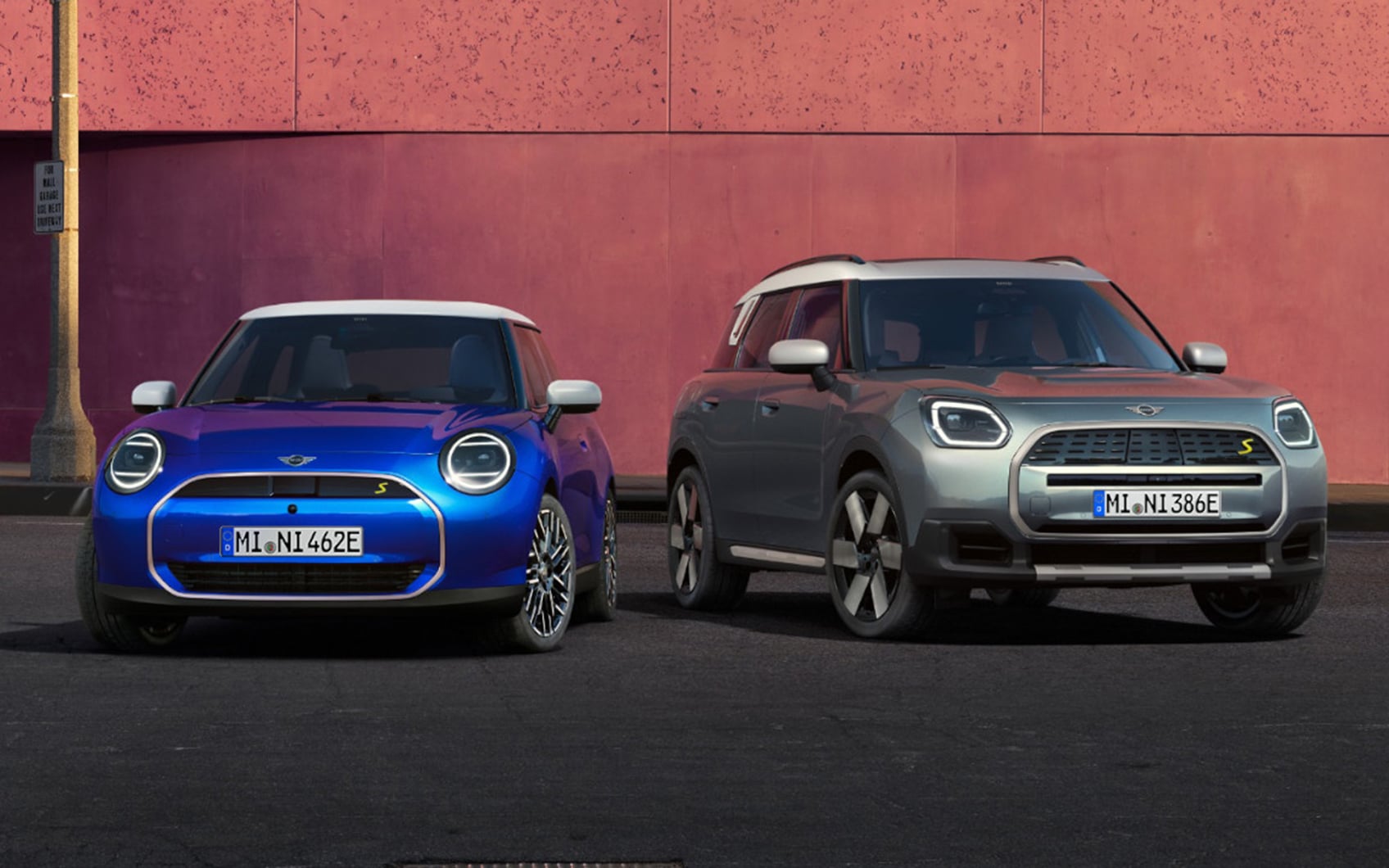 Zwei nebeneinander geparkte MINI-Modelle, ein blauer MINI Cooper SE und ein silberner MINI Countryman SE, vor rosa Wand auf Asphaltfläche.