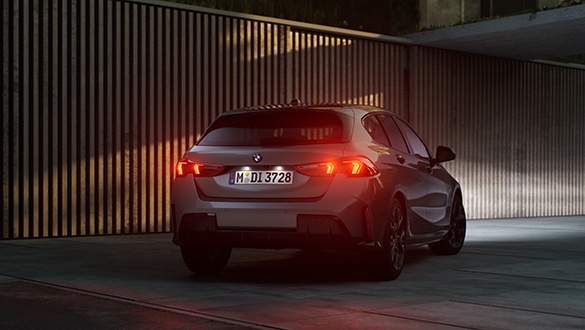 Im Fokus steht das Heck eines grauen BMW 1er. Er parkt in der Dämmerung vor einer Garagenwand und die roten Rückleuchten leuchten.
