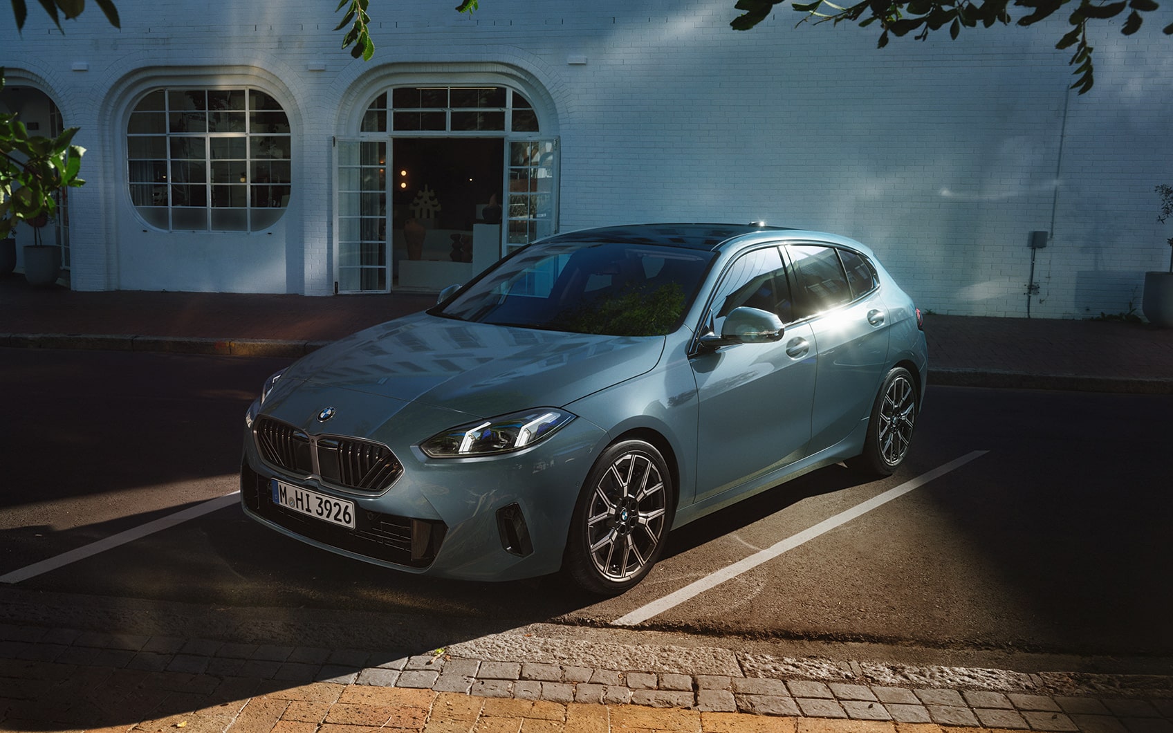 BMW steht auf Hof mit Pflastersteinen. Im Hintergrund ist ein weißes Haus zu sehen. Oben ragen grüne Äste eines Baumes ins Bild.