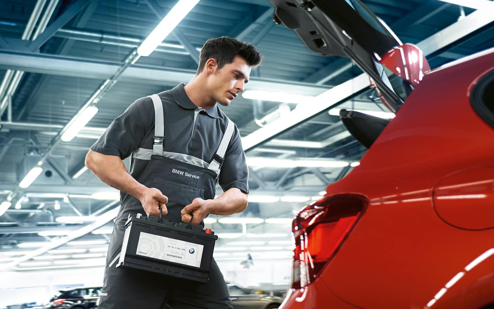 BMW Servicemitarbeiter trägt Autobatterie zu rotem Fahrzeug in Werkstatt.