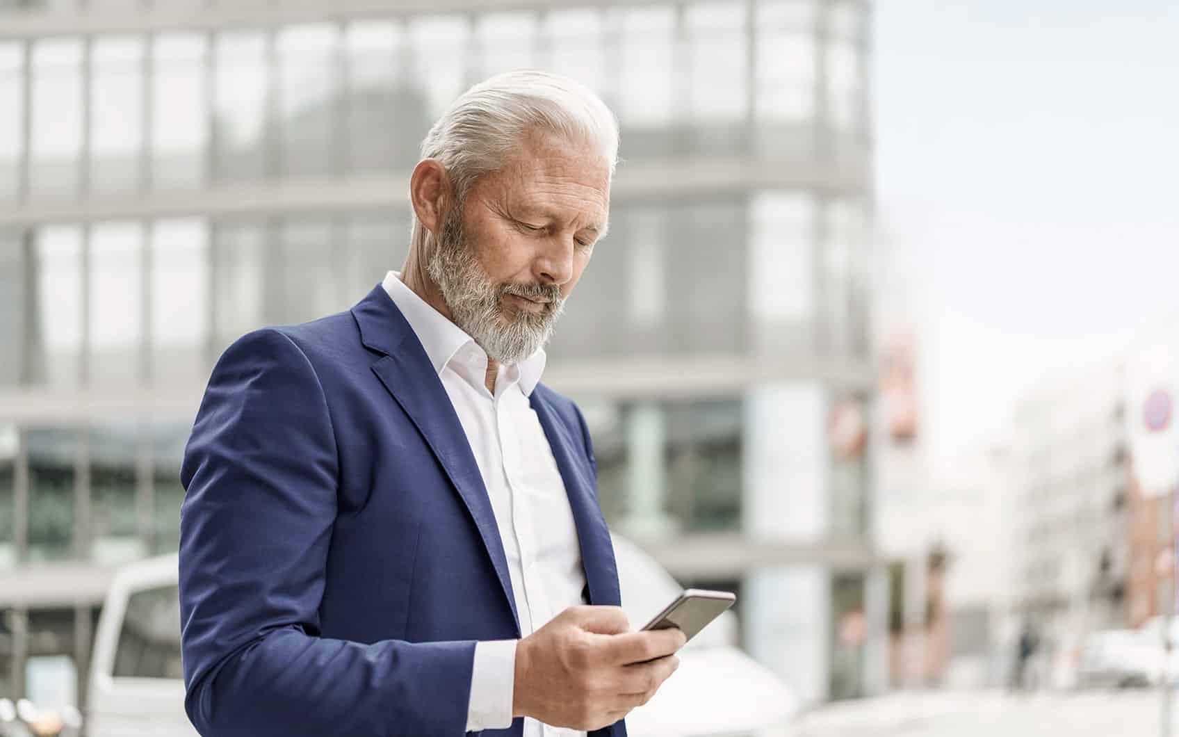 Älterer Mann im Anzug blickt auf Smartphone, moderne Bürogebäude im Hintergrund.