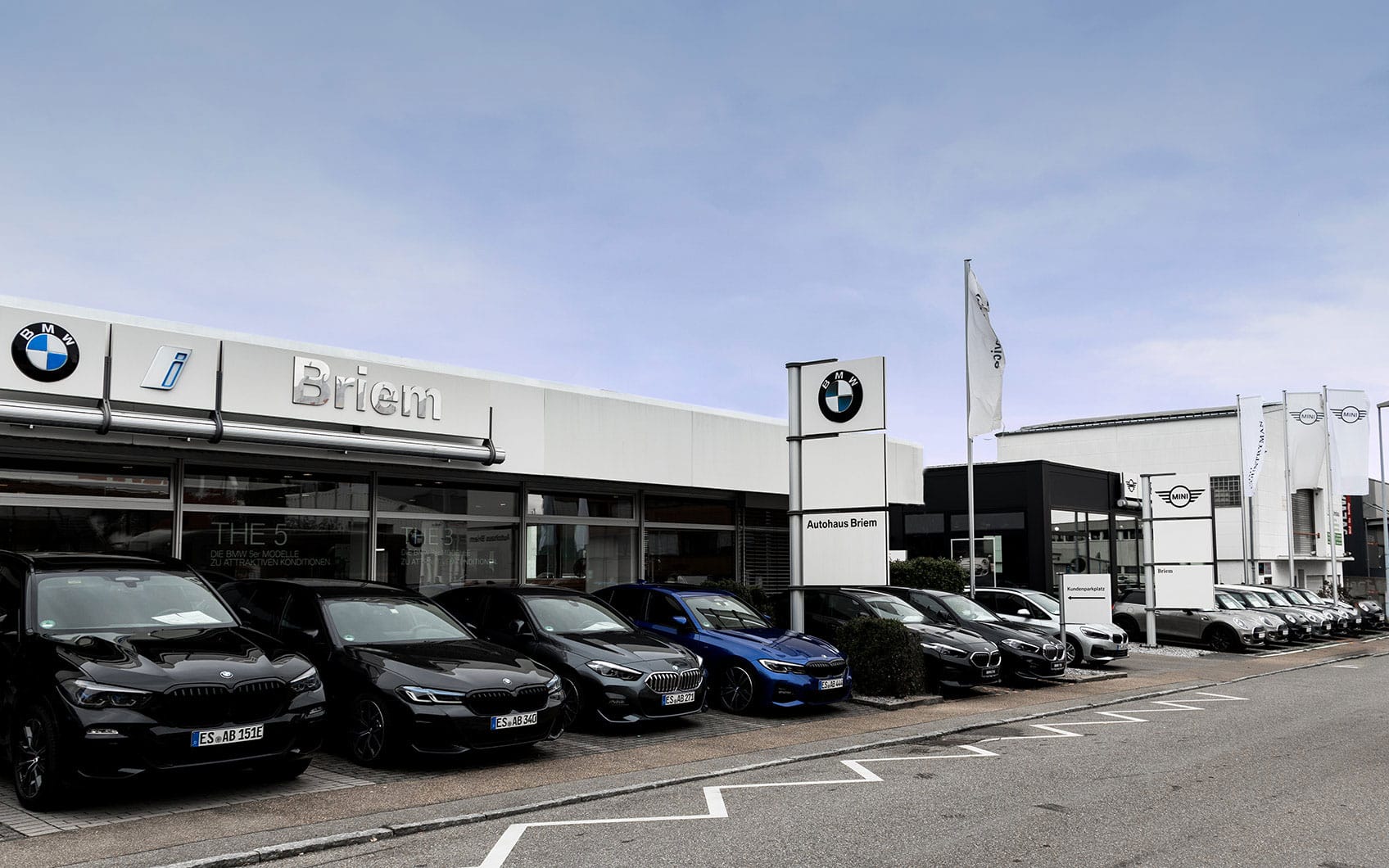 Außenansicht eines BMW-Autohauses mit Verkaufsfläche und parkenden Fahrzeugen vor dem Eingang.