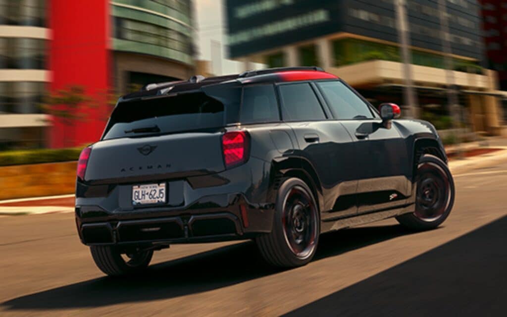 Heckansicht und Profil eines schwarzen MINI John Cooper Works Aceman in Fahrt auf einer Straße mit urbanem Hintergrund