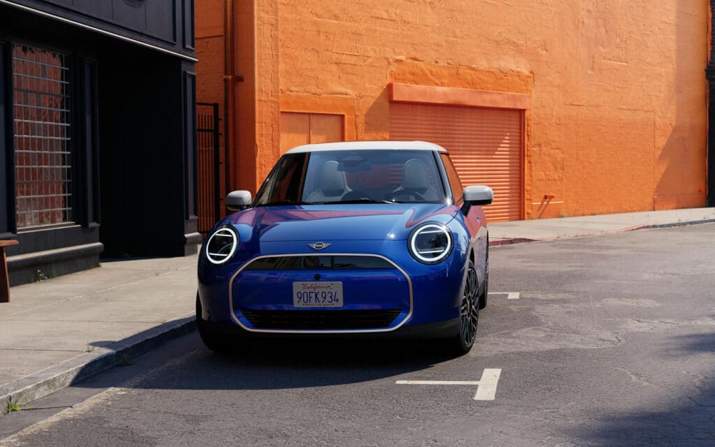 Blauer Mini Cooper Electric mit weißem Dach frontal geparkt vor orangefarbener Wand.