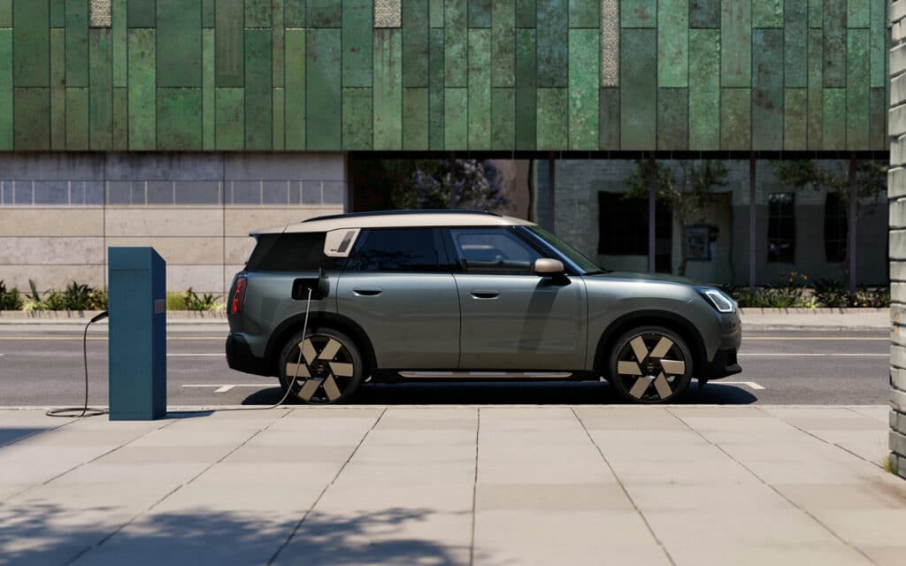 Grüner Mini Countryman Electric mit weißem Dach an Ladesäule auf städtischem Gehweg.