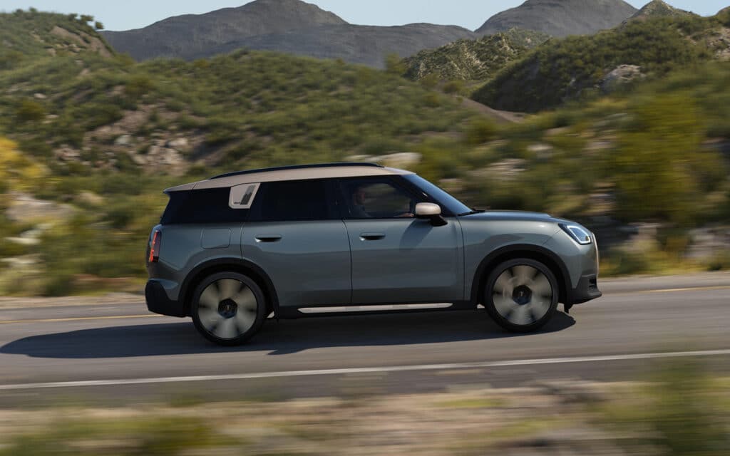 Grüner Mini Countryman mit weißem Dach fährt auf Landstraße vor Bergkulisse.
