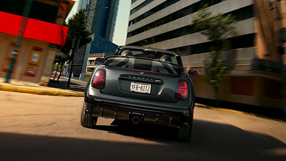 Heckansicht eines fahrenden MINI John Cooper Works Cabrio in der Farbe Schwarz mit Union Jack Heckleuchten und geöffnetem Dach im urbanen Raum