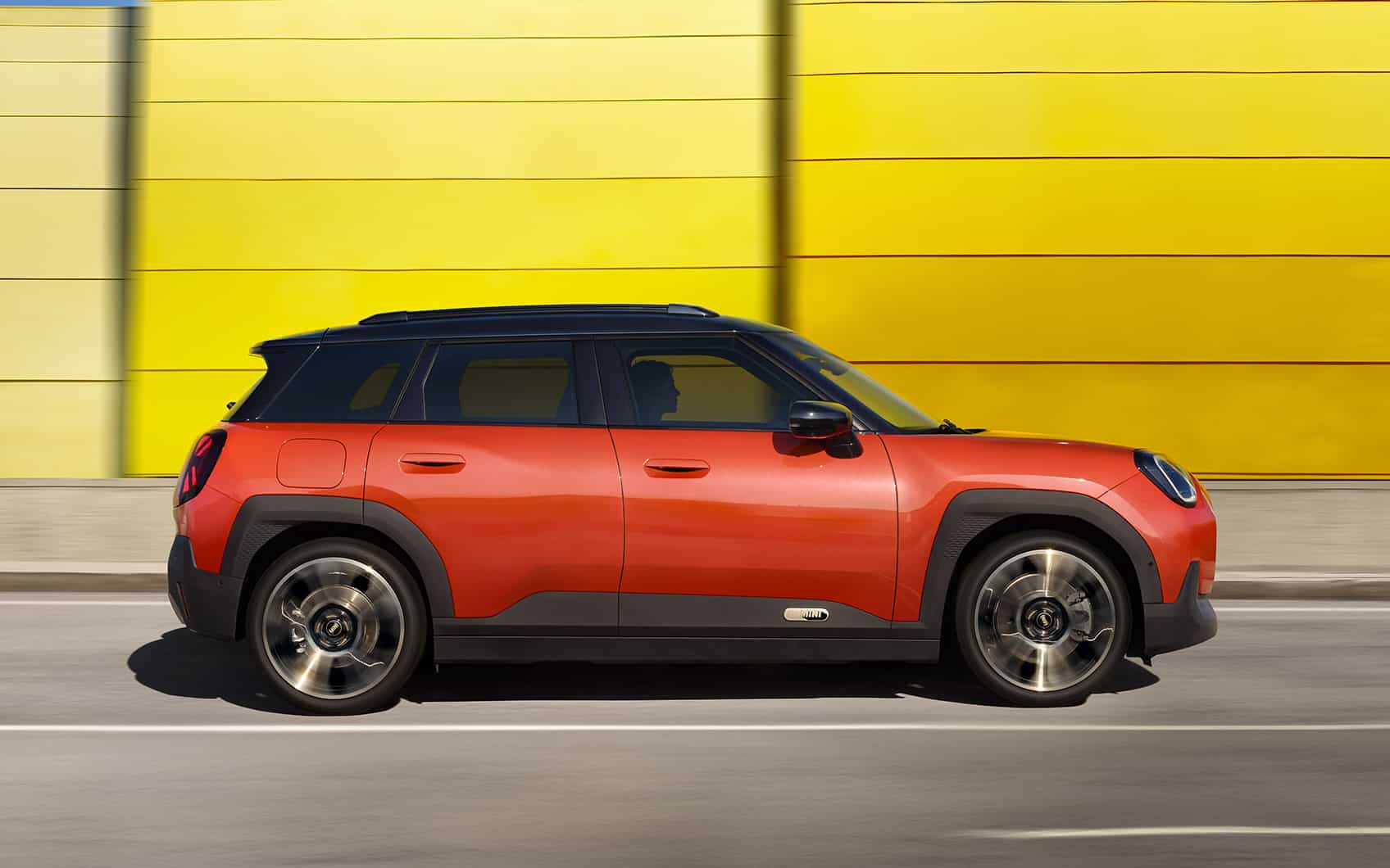 Rotes MINI SUV fährt auf Straße vor gelber Schallschutzwand entlang.