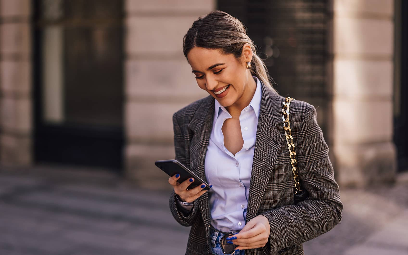 Junge Frau im Business-Outfit schaut lächelnd auf ihr Smartphone beim Gehen durch die Stadt.