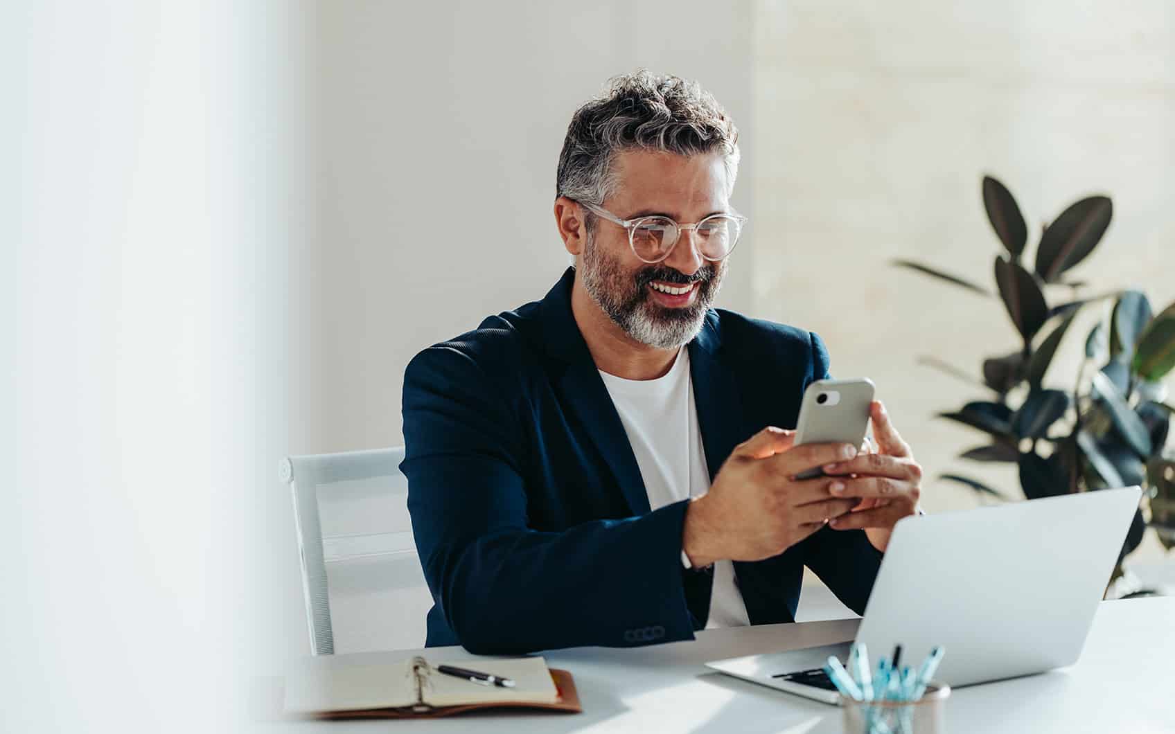 Geschäftsmann mit Brille sitzt am Schreibtisch, lächelt und tippt auf Smartphone.