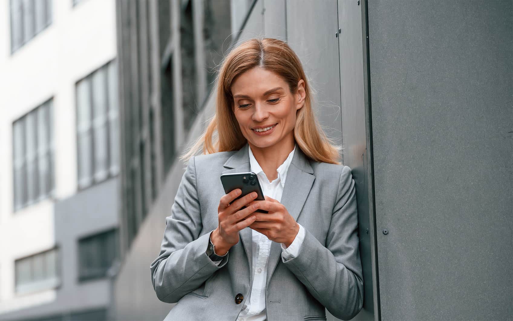 Geschäftsfrau im grauen Anzug lehnt an Wand und tippt lächelnd auf Smartphone.