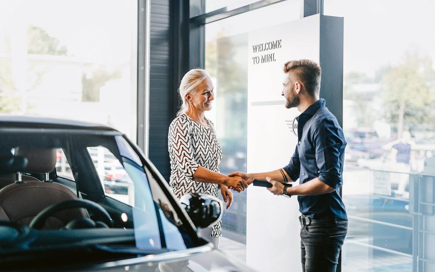 Kundin und Servicemitarbeiter geben sich im Autohaus vor MINI-Schild die Hand.