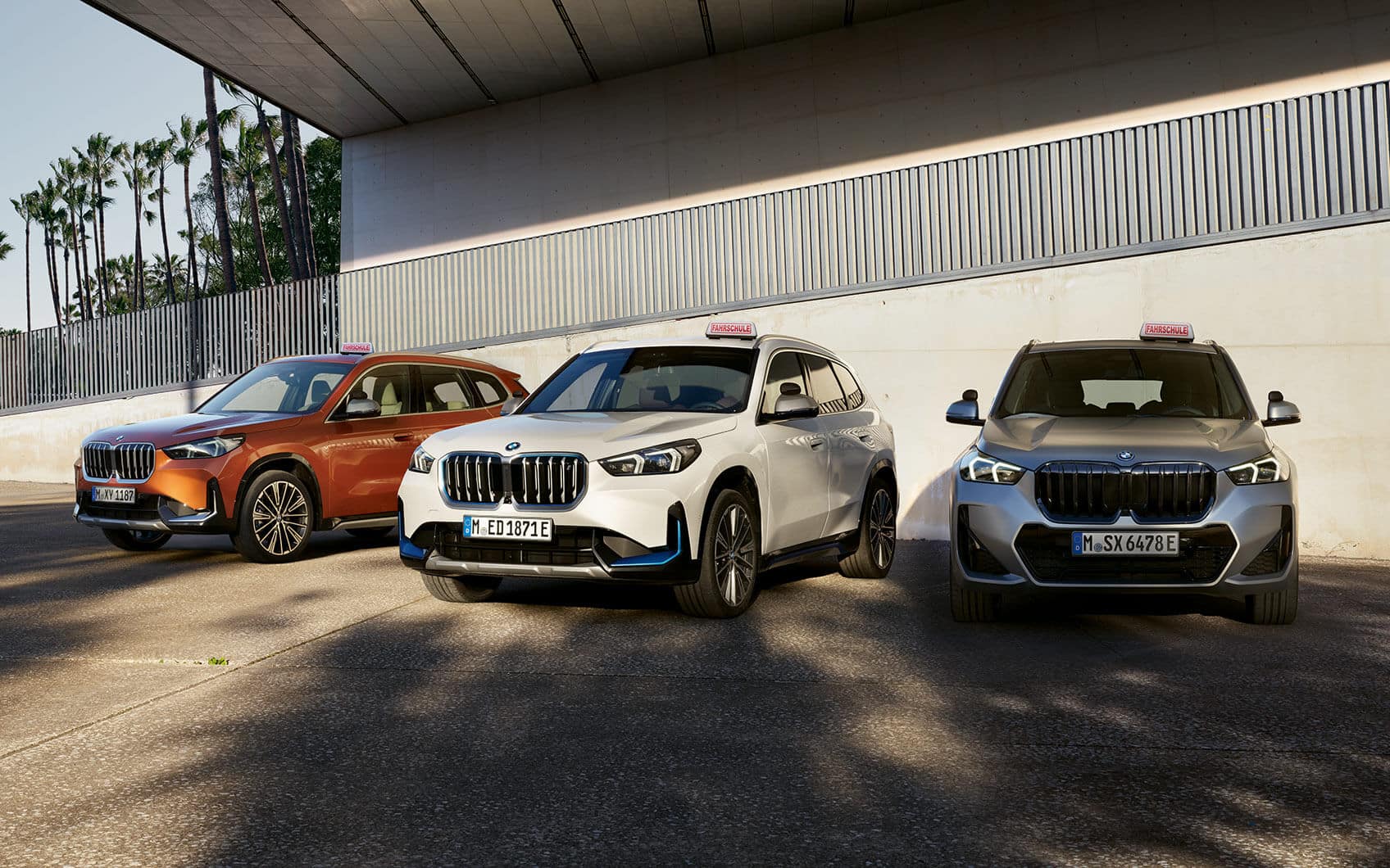 Drei BMW-SUVs mit Fahrschulschildern nebeneinander auf offenem Platz vor moderner Betonwand geparkt.