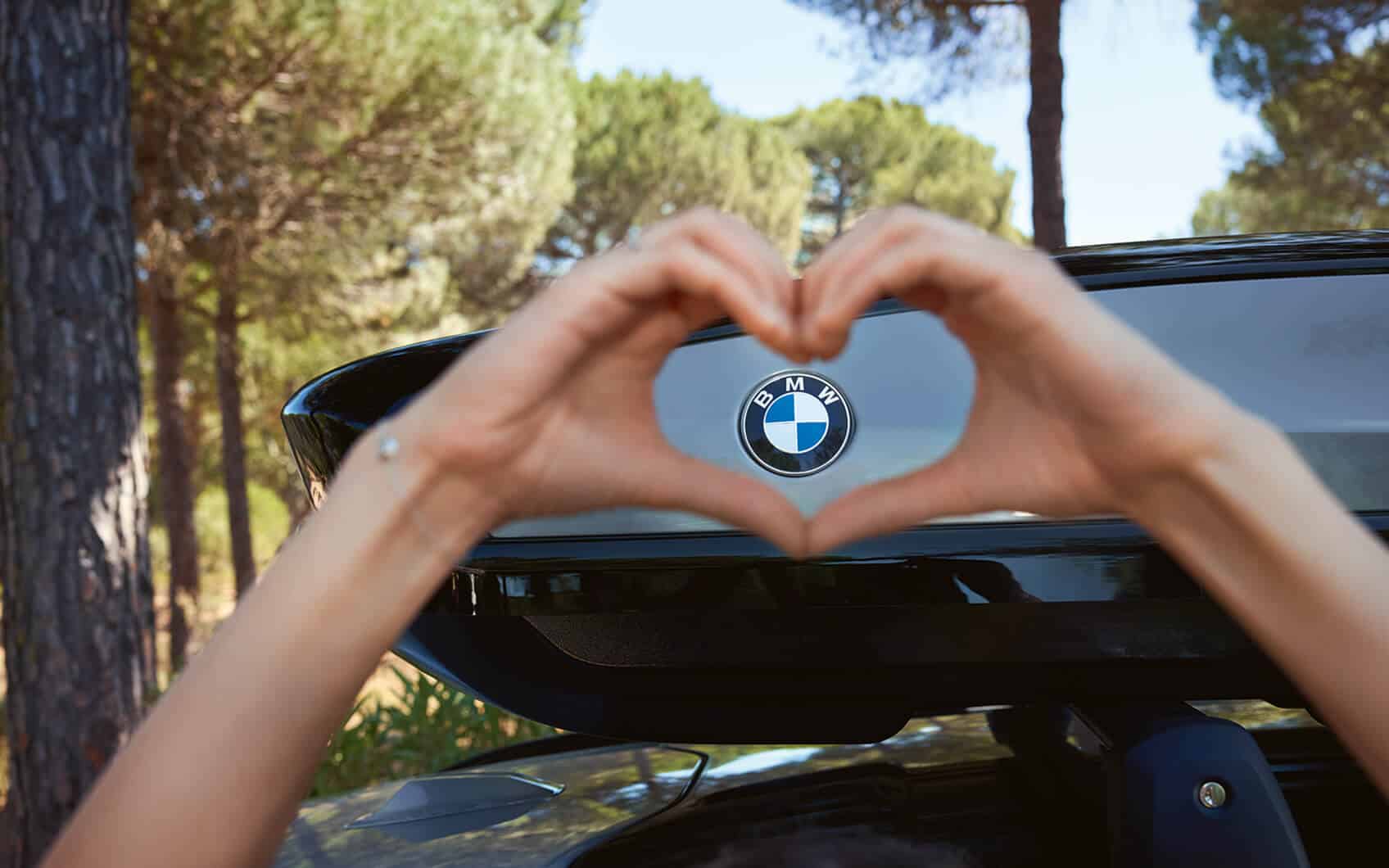 Hände formen ein Herz vor dem BMW-Logo an einem Autoheck im Wald.