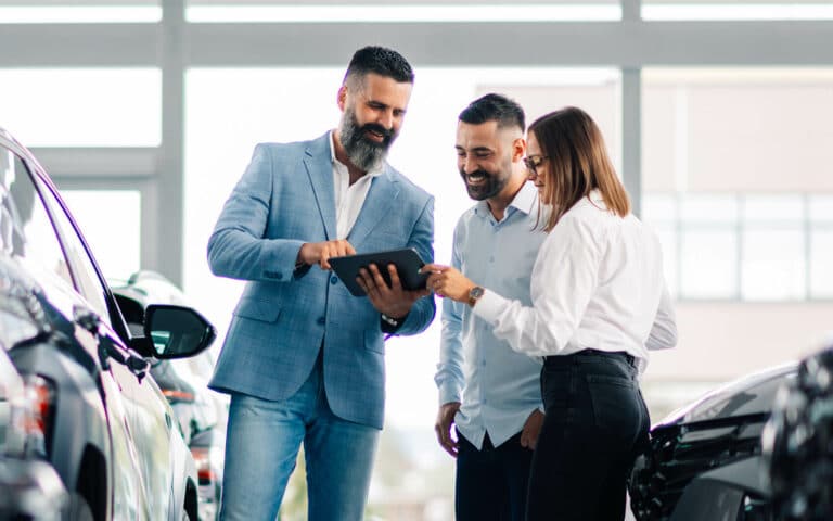 Drei Personen in einem Autohaus betrachten gemeinsam ein Tablet zwischen geparkten Autos.
