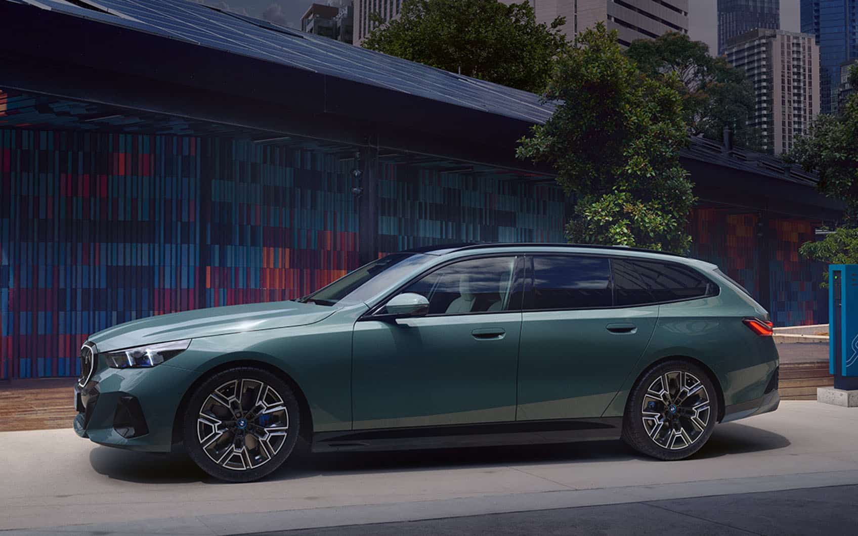 BMW i5 Touring an Ladesäule vor moderner Stadtarchitektur