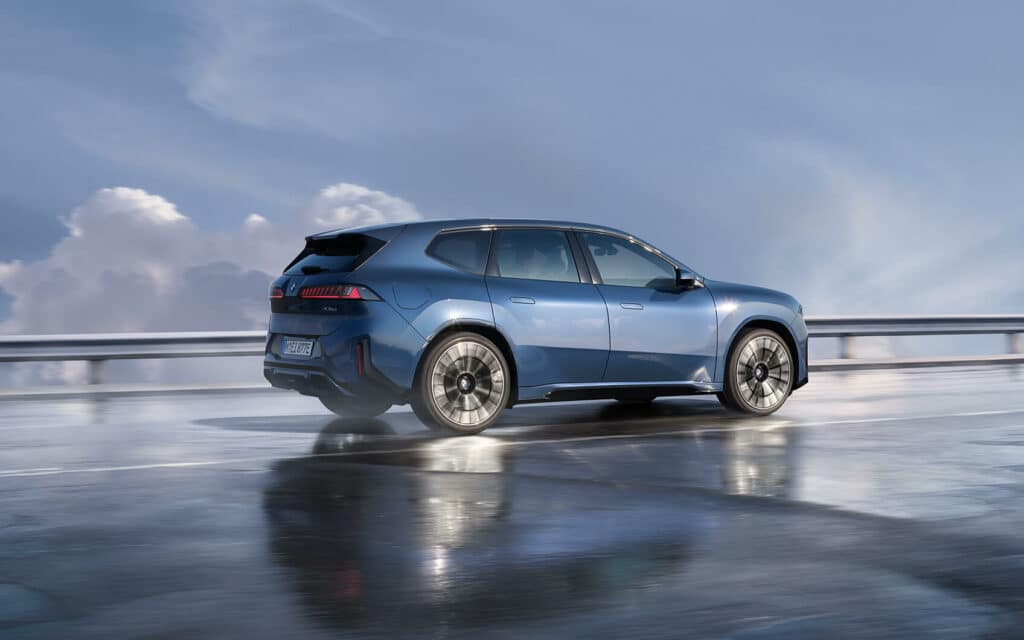 Blauer BMW iX3 fährt bei nasser Straße und bewölktem Himmel auf einer Autobahn.