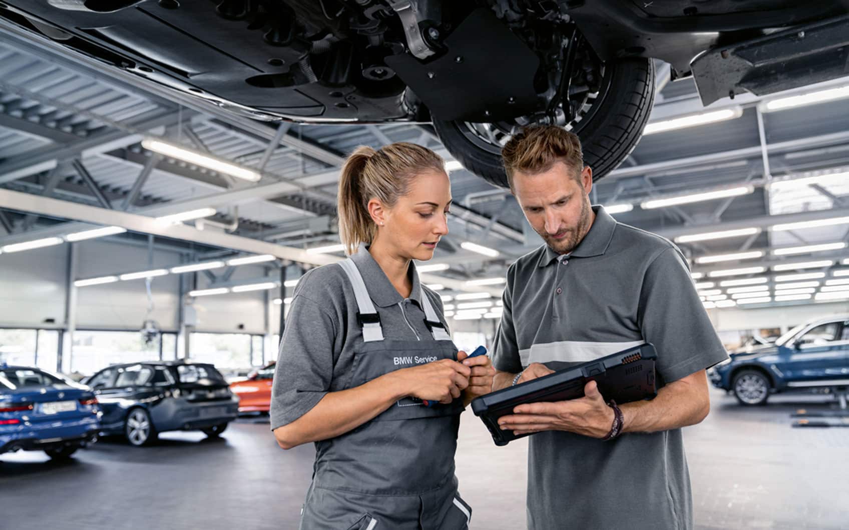 Zwei Kfz-Mitarbeitende in BMW-Werkstatt besprechen Reparaturdaten auf einem Tablet unter einem angehobenen Fahrzeug