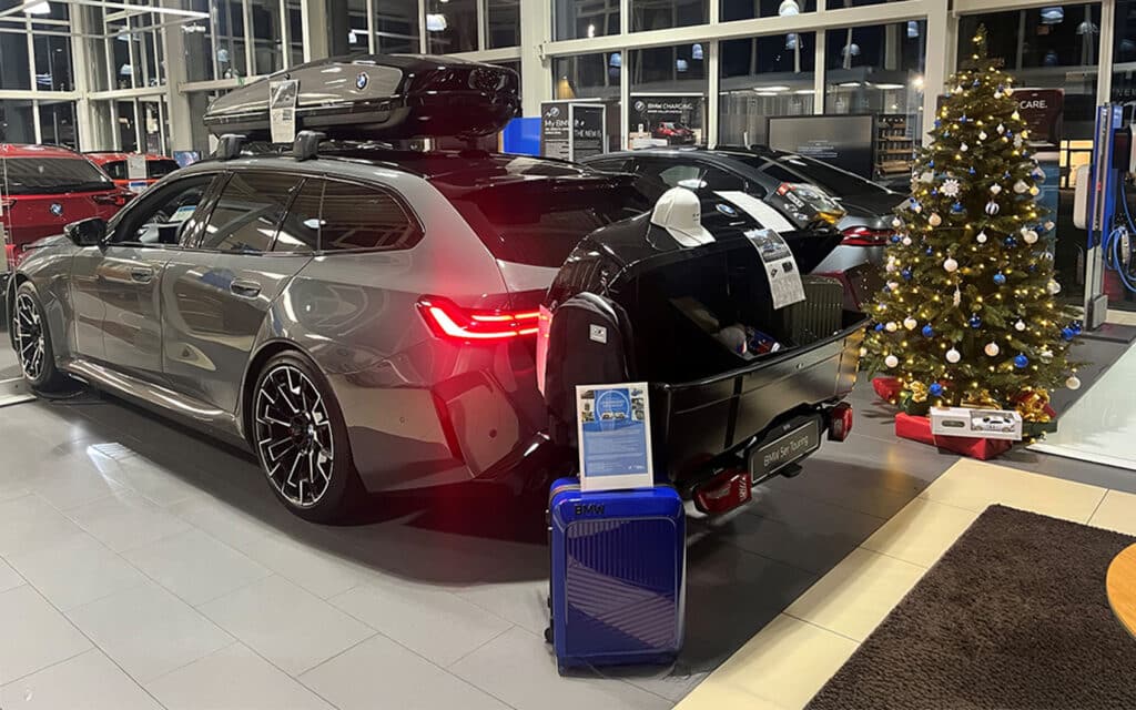 BMW 5er Touring mit Dachbox, Anhängerbox und Gepäck im Autohaus, daneben geschmückter Weihnachtsbaum mit Geschenken