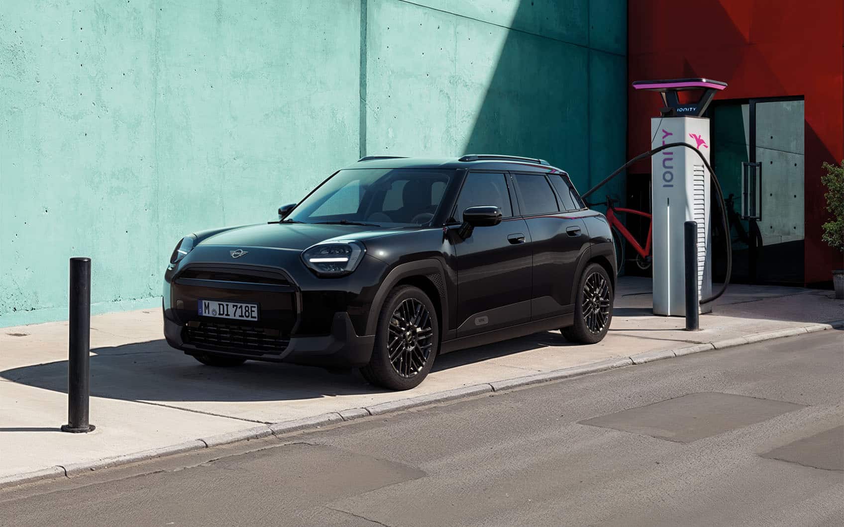 MINI Aceman Blackyard an Ionity-Ladesäule angeschlossen vor türkis-roter Hauswand.