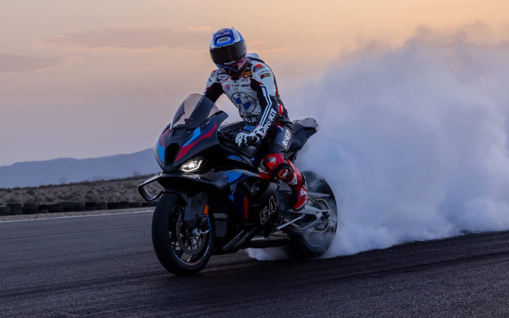 Motorradfahrer in weiß-schwarzer Motorradkleidung mit roten und blauen Akzenten. Er fährt das Motorrad auf einer Straße, hinter dem Motorrad weht eine Dampfwolke