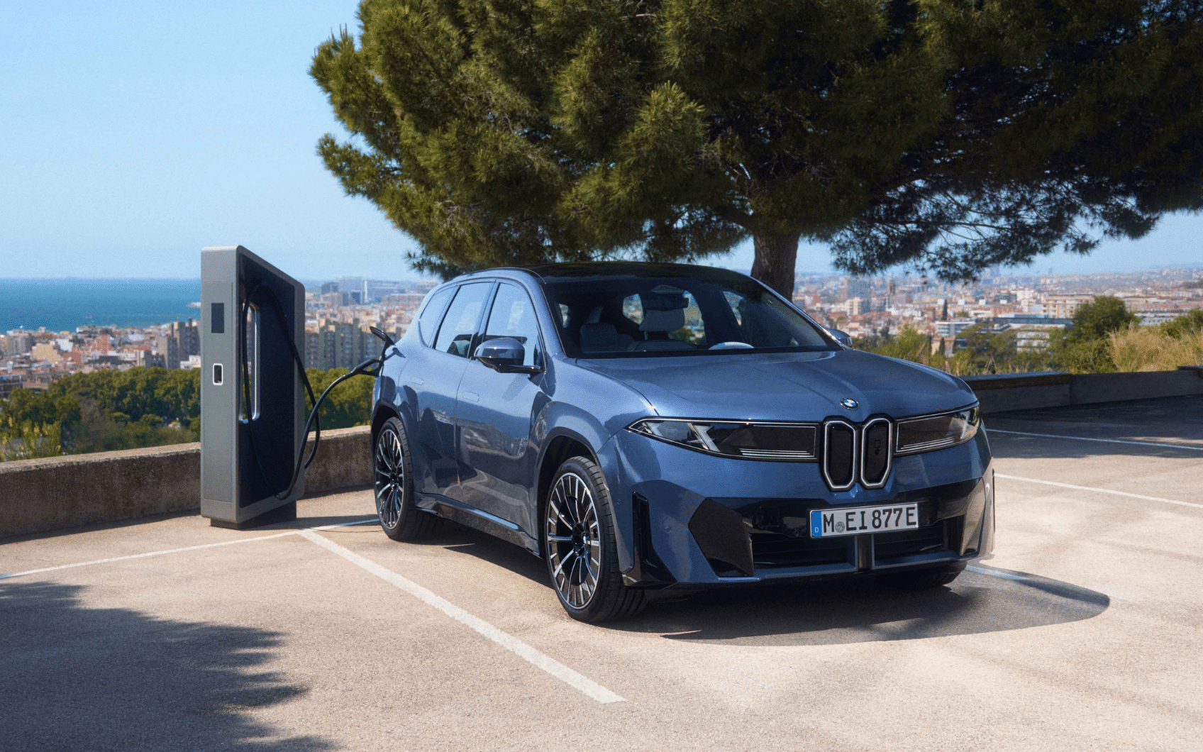 Blauer BMW iX3 beim Laden an einer Ladesäule mit Stadt- und Meerblick im Hintergrund.