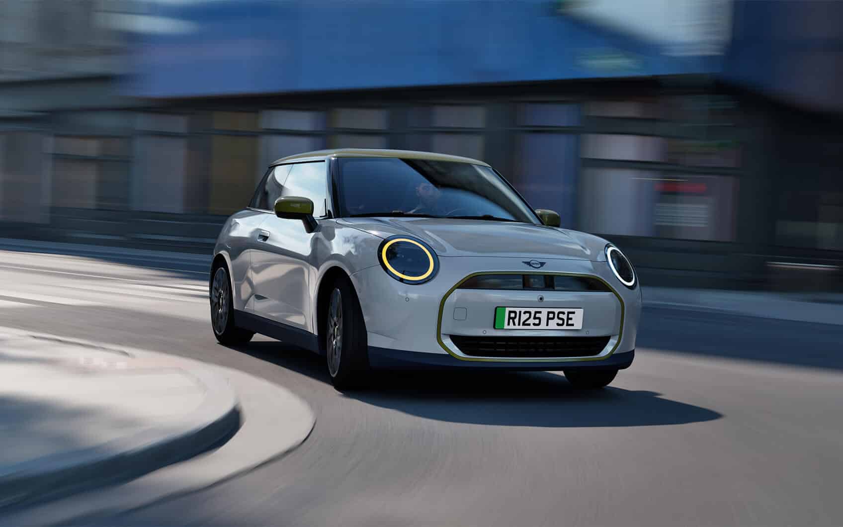 MINI Cooper Electric Paul Smith Edition mit hellem Exterieur und grünem Dach fährt durch eine moderne Stadtstraße.