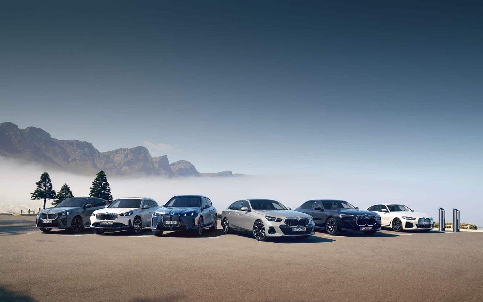 Sechs BMW-Modelle nebeneinander auf einem Parkplatz vor nebelverhüllter Bergkulisse