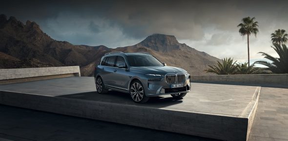 BMW X7 in Grauton auf Betonfläche vor Gebirgspanorama, Frontansicht zeigt Designelemente, in dramatischem Licht